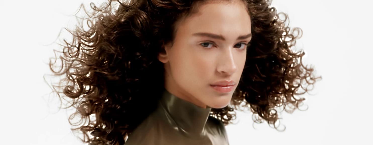 Mujer posando y luciendo su cabello rizado sano, radiante y bien definido, con volumen, definición y brillo natural.