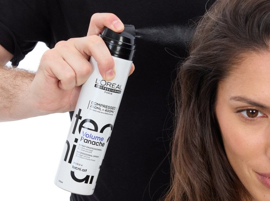El foco se centra en el cabello largo de una mujer que gana volumen mientras un estilista profesional utiliza un spray voluminizador, siguiendo la última tendencia en peinados.